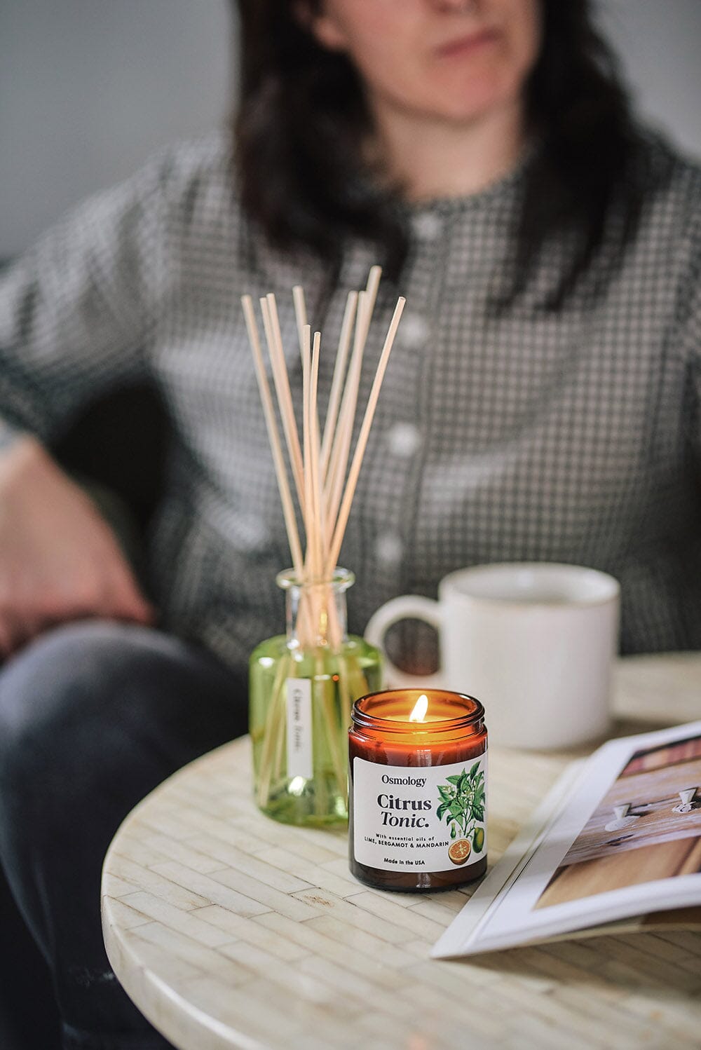 Osmology Citrus Tonic jar candle with lit candle flame on coffee table, Citrus Tonic diffuser behind with a mug, woman sat in background