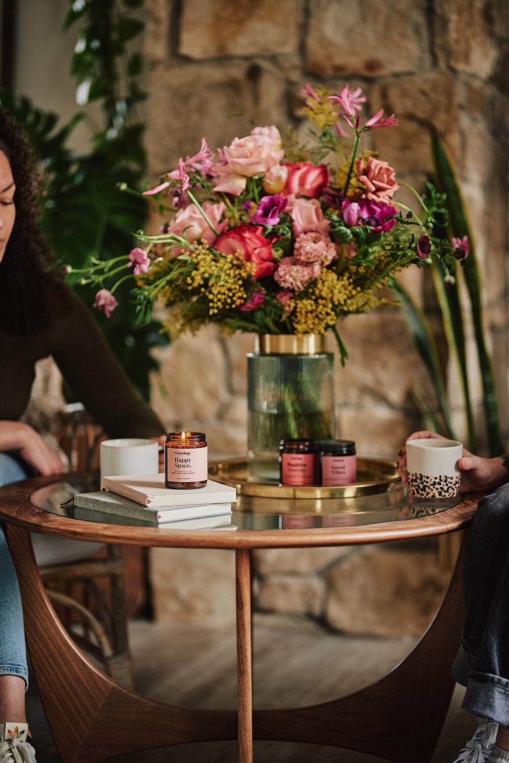 A coffee table with two people sat at, Osmology Happy Space jar candle with lit candle flame placed on books, colourful flowers in vase on table, two other jar candles on table behind