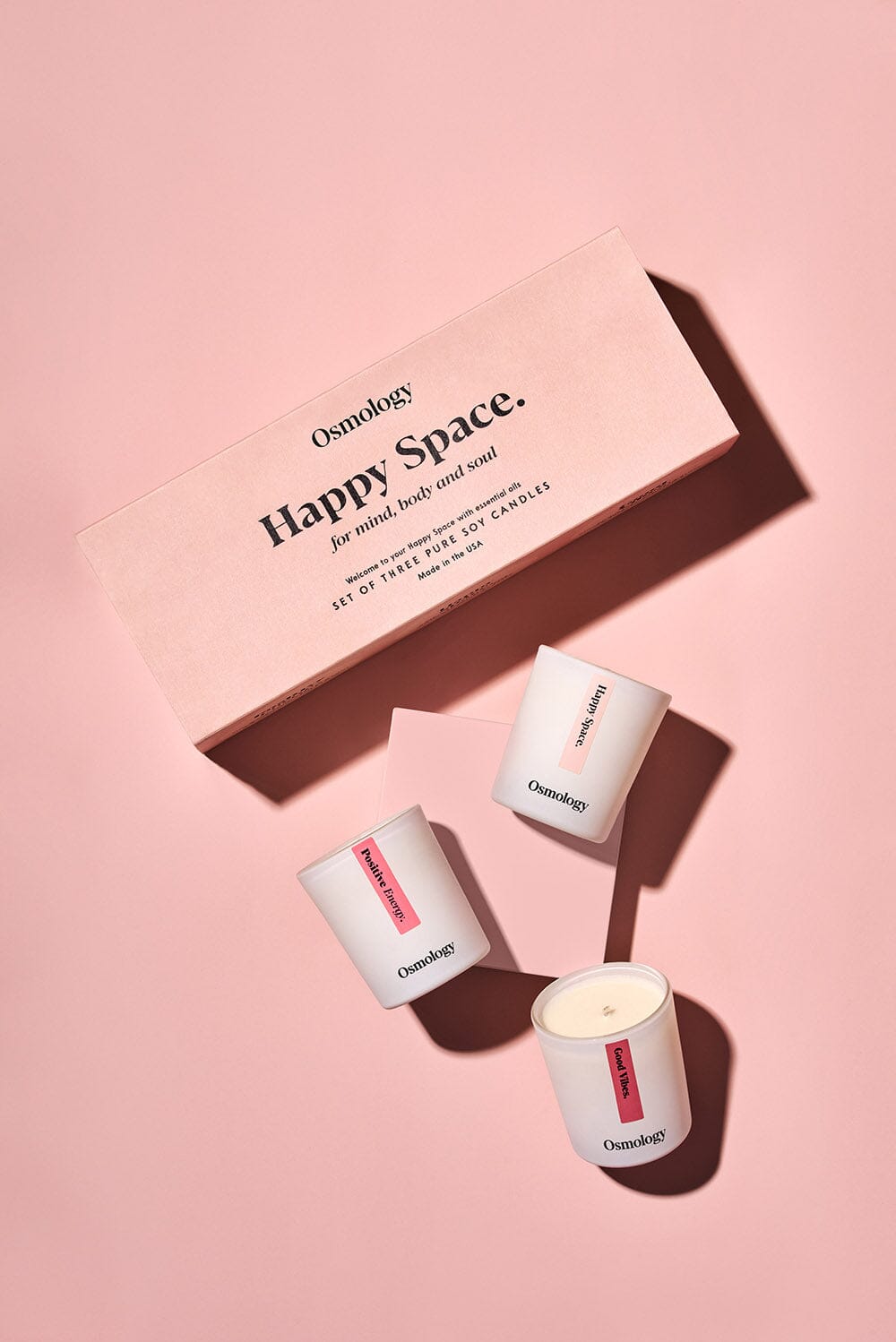 Osmology Happy Space Gift Set, three mini candles including Positive Energy, Happy Space and Good Vibes arranged around a square plinth, product packaging above, flat on foor, pink background