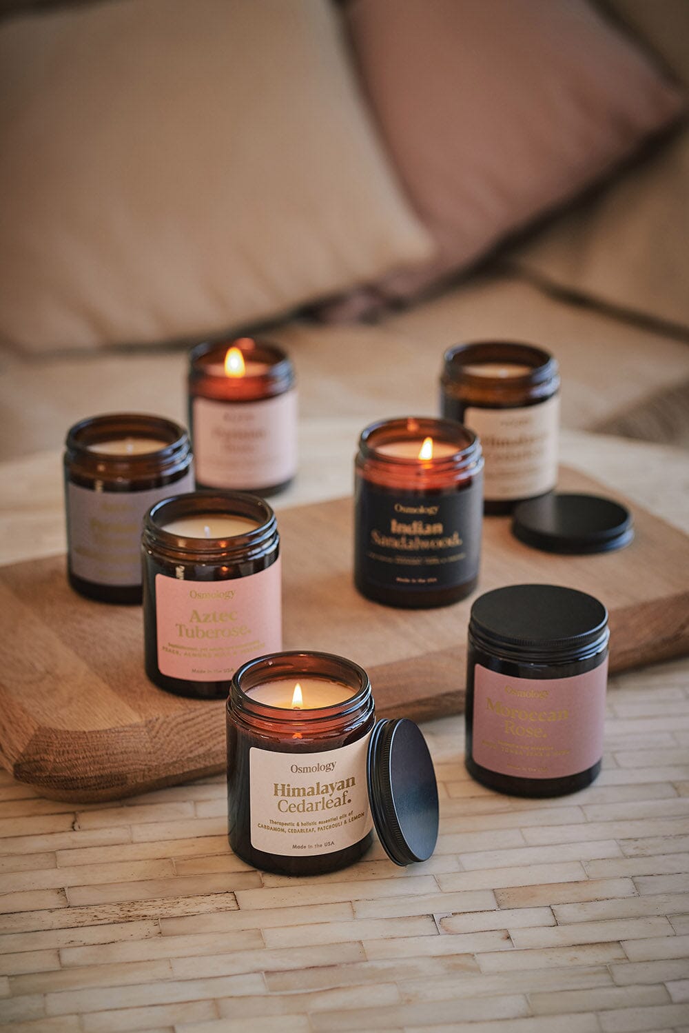 A range of Osmology Fernweh jar candles arranged on table and wooden board, Himalyan Cedarleaf jar candle in foreground with lit candle flame with lid leaning against it