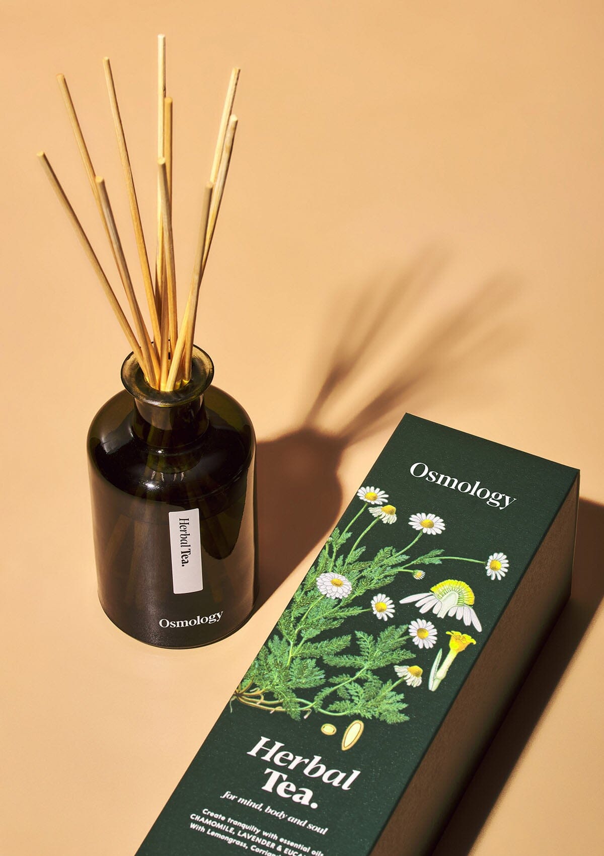 Osmology Herbal Tea reed diffuser in dark green glass next to product packaging on an orange-cream background