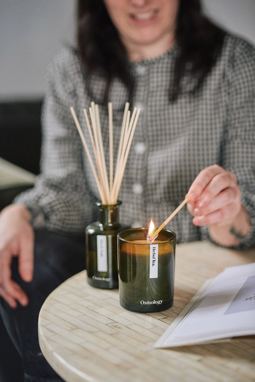 Osmology Herbal Tea candle sat on coffee table being lit by person from behind, reed diffuser placed behind candle