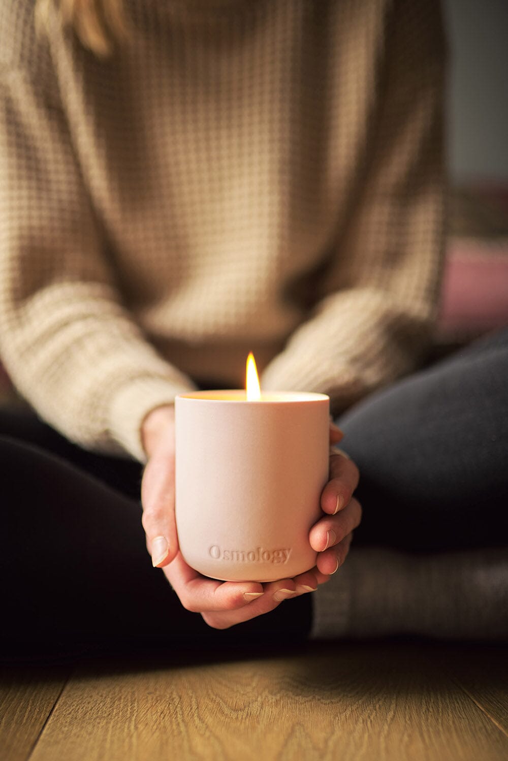 Osmology Himalayan Cedarleaf candle with lit flame being held with two hands, a girl sat cross legged on the floor