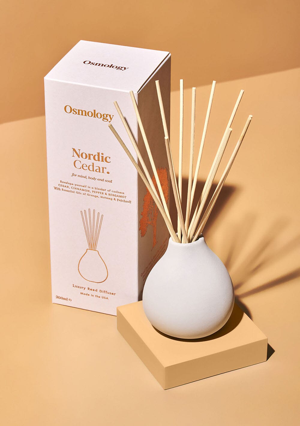 Osmology Nordic cedar ceramic reed diffuser in white next to product packaging on an orange-cream background