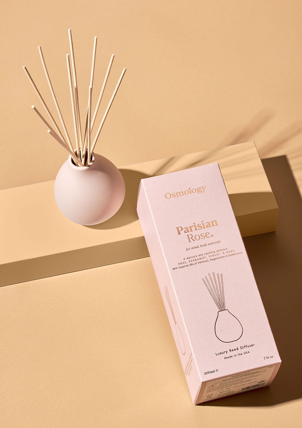 Osmology Parisian Rose pale pink reed diffuser next to product packaging on an orange background