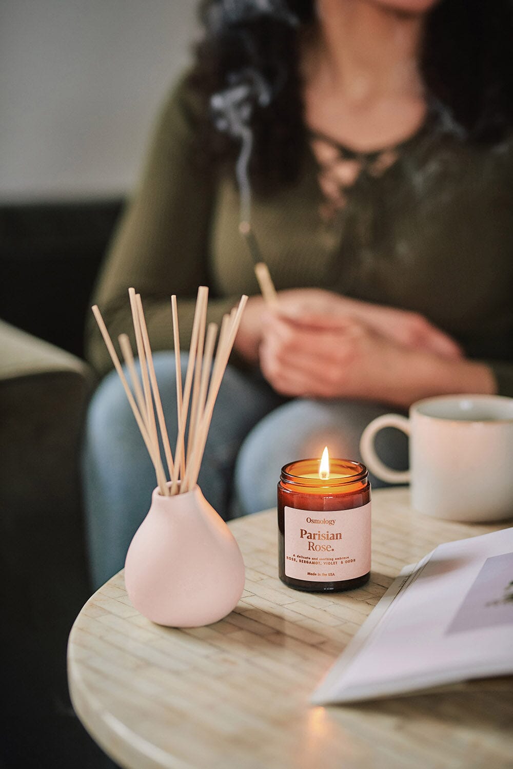 Osmology Parisian Rose jar candle with lit flame and reed diffuser placed on table